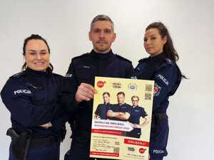 Zdjęcie kolorowe. Troje policjantów pozujących do zdjęcia. Wszyscy w umundurowaniu koloru granatowego. Policjant stojący w środku trzyma plakat promujący konkurs Super Dzielnicowy 2026