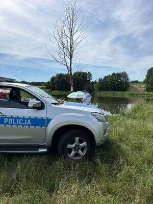 Zdjęcie boku policyjnego radiowozu, który stoi na trawie. Przed radiowozem, z drugiej strony stoi policjant.