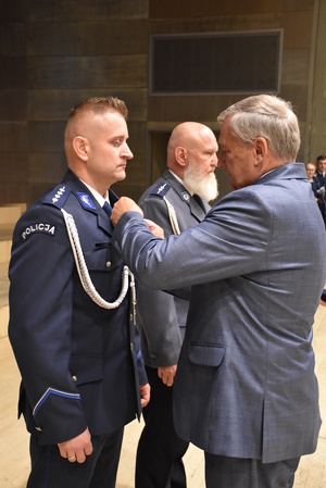 Minister przypina policjantowi do munduru odznaczenie. Obok stoi drugi policjant.