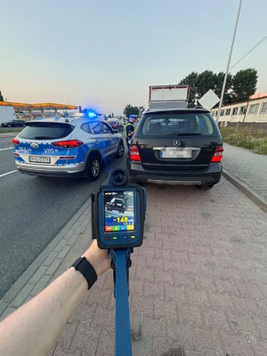 Zdjęcie kolorowe. Policyjny oznakowany radiowóz stoi obok pojazdu marki Mercedes koloru czarnego. Po miedzy pojazdami stoi policjant. Zdjęcie wykonane jest od tyłu stojących pojazdów. na pieszym planie widać rękę, która trzyma urządzenie do mierzenia prędkości, na którym widać zapis pomiaru -148 km/h.