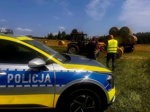 Zdjęcie kolorowe. Na pierwszym planie fragment radiowozu. Za nim policjantka i przedstawiciel Państwowej Inspekcji Pracy rozmawiają z rolnikiem.  W tle widać ciągnik i załadowane na przyczepie baloty ze słomy.