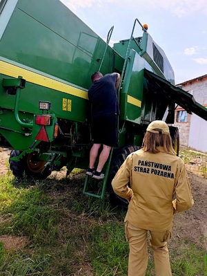 Na pierwszym planie funkcjonariusz Państwowej Straży Pożarnej odwrócony tyłem. Stoi przed kombajnem na schodkach którego stoi rolnik.
