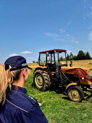 Zdjęcie kolorowe. Policjantka stoi obok ciągnika rolniczego zaparkowanego na łące.