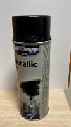 Zdjęcie kolorowe. Szara puszka, spray na farbę z czarnym korkiem. Napisy i grafiki na puszcze zostały zapikselowane.