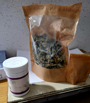 Zdjece kolorowe. Od lewej strony opakowanie plastikowe koloru białego po lekach z zapikselowanymi napisami. Obok duże opakowanie koloru brązowego z przeźroczystym okienkiem, w którym widać susz koloru zielonego.
