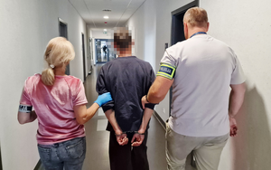 Zdjęcie kolorowe. Dwójka nieumundurowanych policjantów prowadzi zatrzymanego, który ma ręce skute kajdankami. Zdjęcie robione z tyłu.