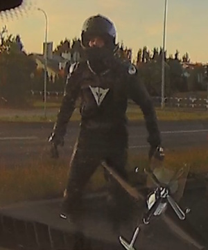 Zrzut z ekranu. Motocyklista w czarnym kominezonie z kaskiem na głowie stoi na jezdni.
