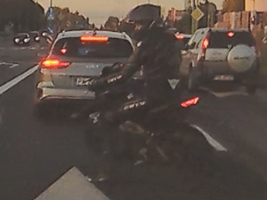 Motocyklista siedzący na motocyklu, ubrany w ciemny kombinezon i kask. Przed nim stojące samochody