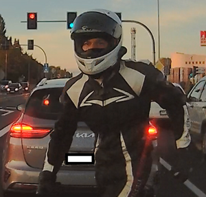 Zrzut z ekranu. Motocyklista w czarno-białym kombinezonie i białym kasku, z podniesioną przesłoną idzie jezdnią. W tle widać stojące pojazdy i sygnalizację świetlną.