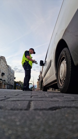 Zdjęcie kolorowe. Zaparkowany pojazd, obok którego stoi funkcjonariusz policji. Zdjęcie robione z dołu.