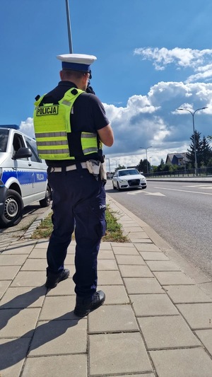Zdjęcie kolorowe. Policjant stojący tyłem przy drodze po której jedzie samochód osobowy koloru białego. Obok, po lewej stronie zdjęcia widać fragment zaparkowanego, oznakowanego radiowozu.
