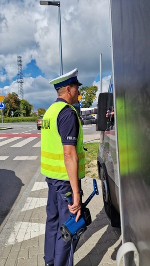 Zdjęcie kolorowe. Policjant stojący obok zaparkowanego pojazdu dostawczego. Policjant trzyma w prawej dłoni urządzenie do pomiaru prędkości.