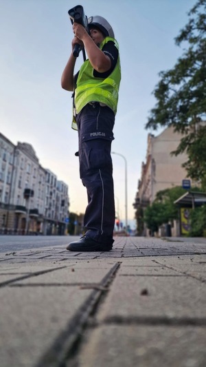 Policjantka stojąc przy krawędzi jezdni mierzy prędkość nadjeżdżających pojazdów. Zdjęcie robione od dołu.