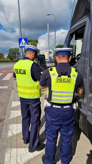 Zdjęcie kolorowe. Dwóch policjantów stoi przy zaparkowanym samochodzie i rozmawia z kierowcą.