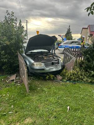 Zdjęcie kolorowe. Auto, które przodem uderzyło w ogrodzenia posesji uszkadzając betonowy płot.  W tle policjant stojący przy radiowozie w którym siedzi zatrzymany kierowca.