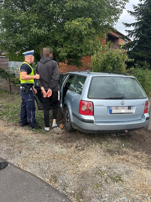 Zdjęcie kolorowe. Policjant z zatrzymanym kierowcą, który ma ręce skute kajdankami z tyłu, stoi przy samochodzie, który uderzył w ogrodzenie posesji. W tle widać drzewo przysłaniające budynek stojący na posesji.