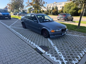 Zdjęcie kolorowe. Miejsce parkingowe oznaczone kopertą, na którym zaparkowany jest pojazd marki BMW. Z tyłu, w oddali za BMW zaparkowany jest policyjny radiowóz. W tle widać fragment parkingu z zaparkowanymi pojazdami i drogę.