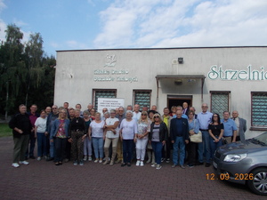 Zdjęci kolorowe. Grupa osób pozujących do zdjęcia, stojąc przed budynkiem z napisem Kaliskie Bractwo Strzelców Kurkowych Strzelnica. W prawym dolnym rogu widać fragment zaparkowanego auta.